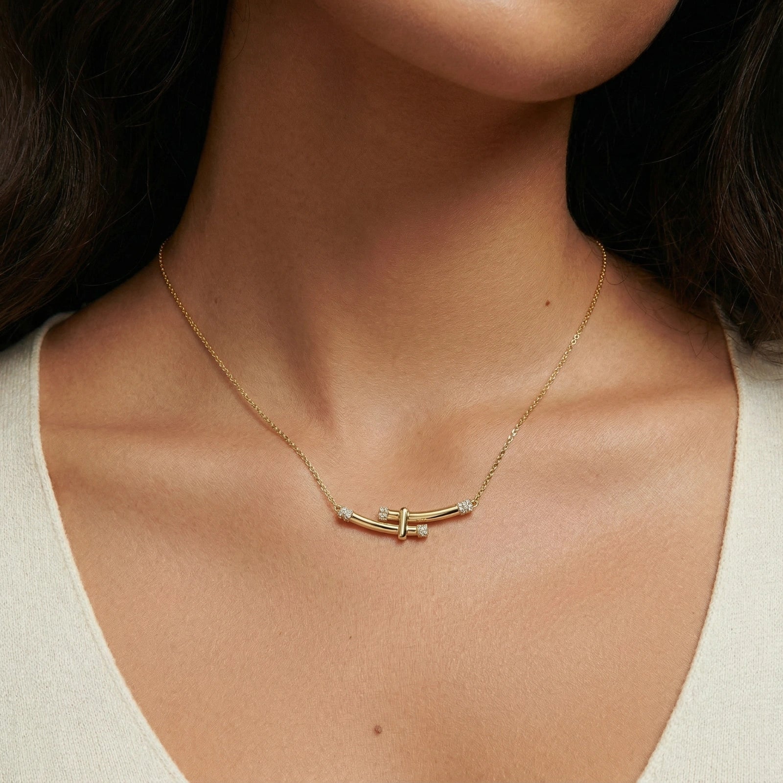 Close-up of a woman's neck wearing a delicate gold necklace with a curved bar design and small diamonds. She is wearing a light-colored top.
