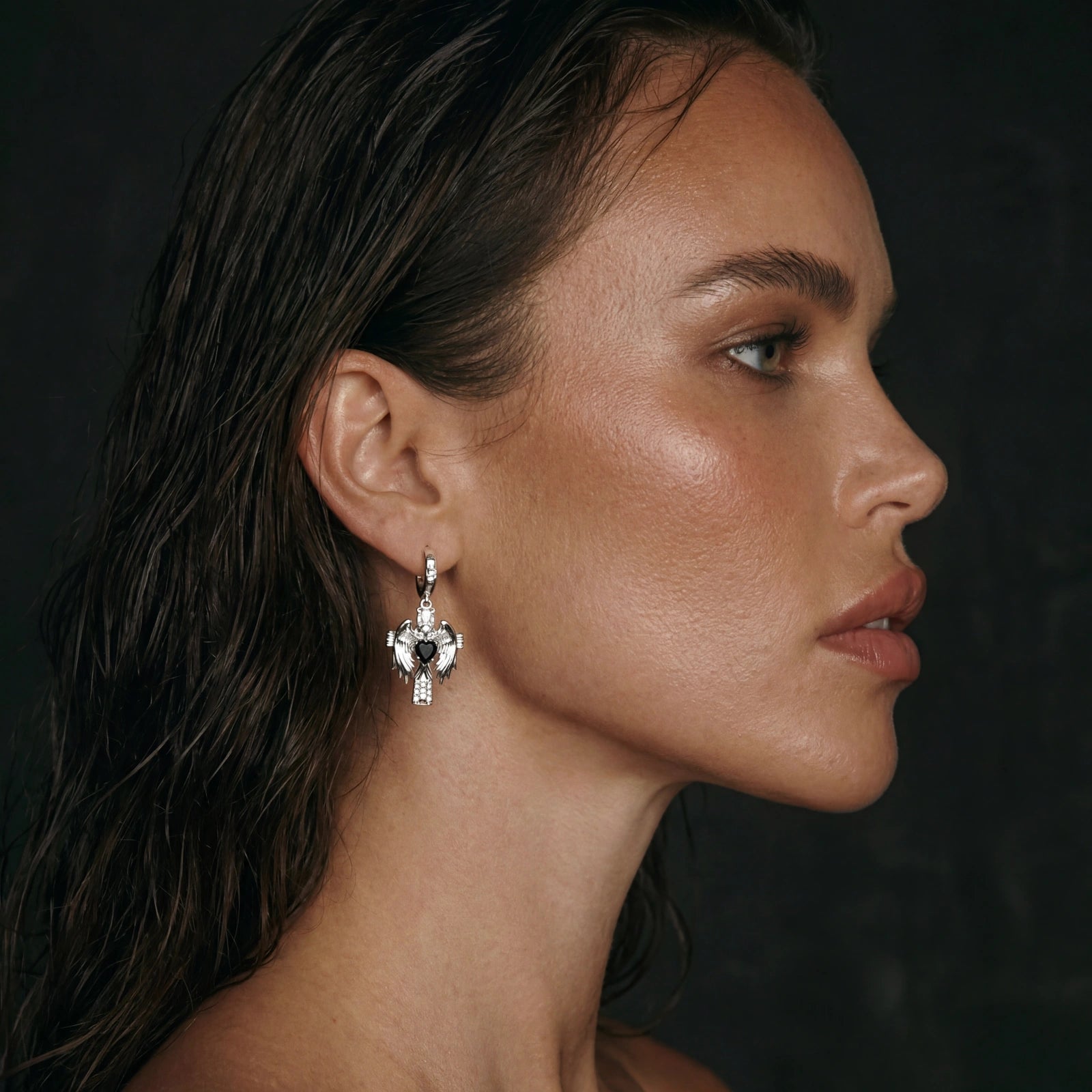 A woman with wet hair is shown in profile against a dark background. She is wearing a large, ornate silver earring shaped like an abstract bird.